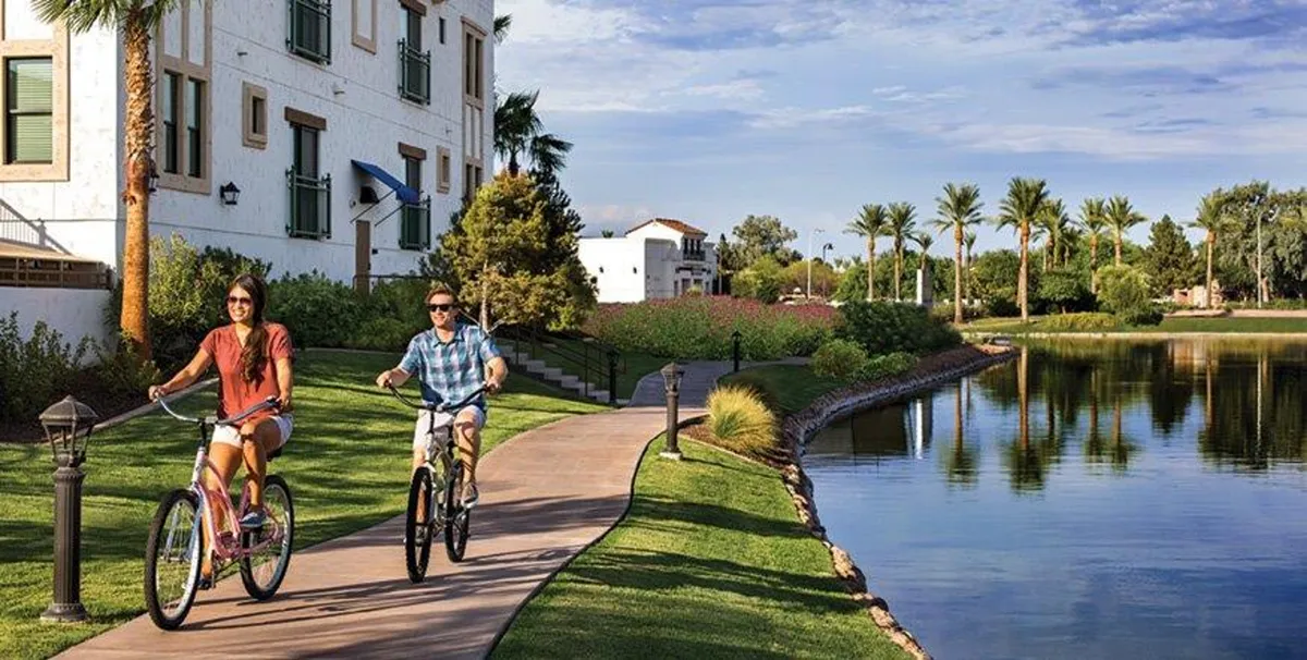 This exterior image highlights two bicyclists riding along a scenic lakeside pathway, framed by palm trees and water, with a nearby building in the background. The vibrant greenery, blue sky, and relaxed activity capture the spirit of outdoor exploration and leisure that appeals to golf lovers seeking a resort-like atmosphere.