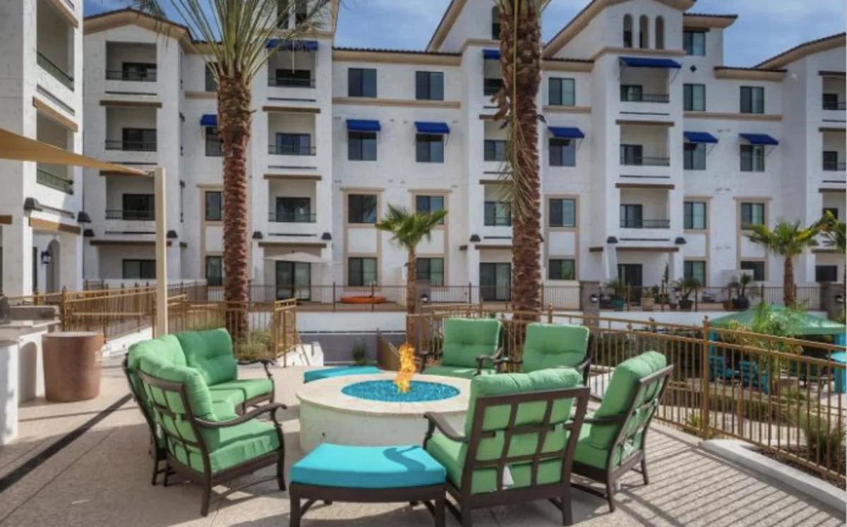 A welcoming outdoor fire pit area with bright green armchairs arranged for conversation, surrounded by palm trees and blue-accented architecture. The setting suggests cozy evenings and daytime gatherings in a vibrant, friendly community environment.