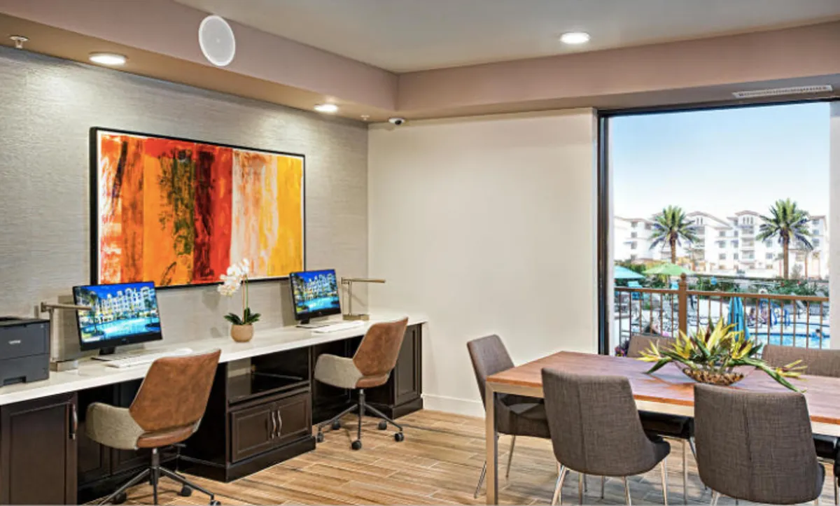 A stylish business center featuring computers, colorful artwork, a dining table adorned with plants, and a relaxed yet productive atmosphere. The space supports both collaborative meetings and solo work sessions.