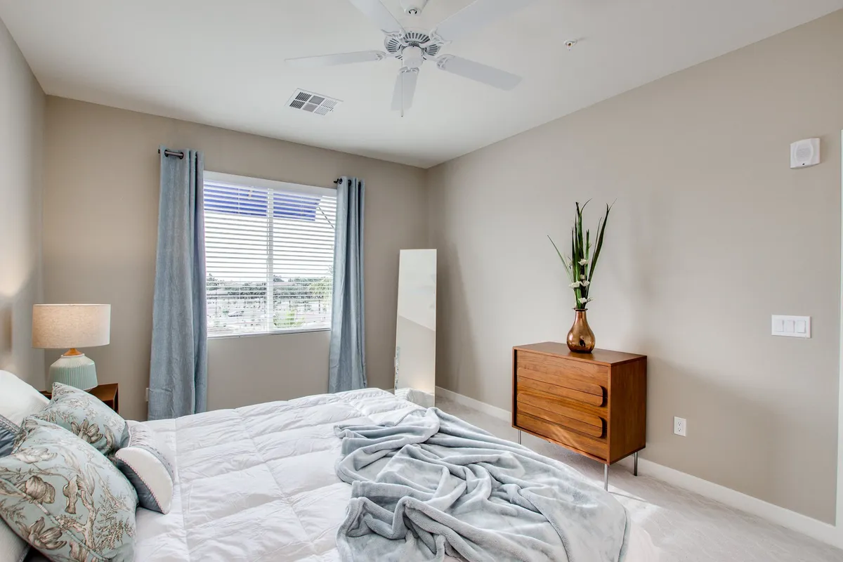 A cozy and serene bedroom with decorative pillows on the bed, a wooden dresser, a large window with curtains, a floor mirror, and an indoor plant. The overall atmosphere is inviting, offering plenty of natural light and space to relax or get ready for the day.