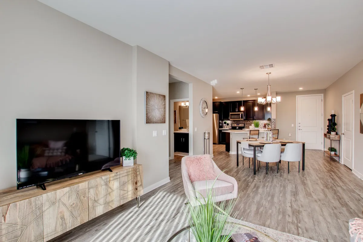 A cozy living area featuring a television on a wooden console, adjacent dining table with chairs, and a glimpse into the kitchen. Decorative touches like a potted plant and warm lighting create a welcoming atmosphere ideal for shared activities.