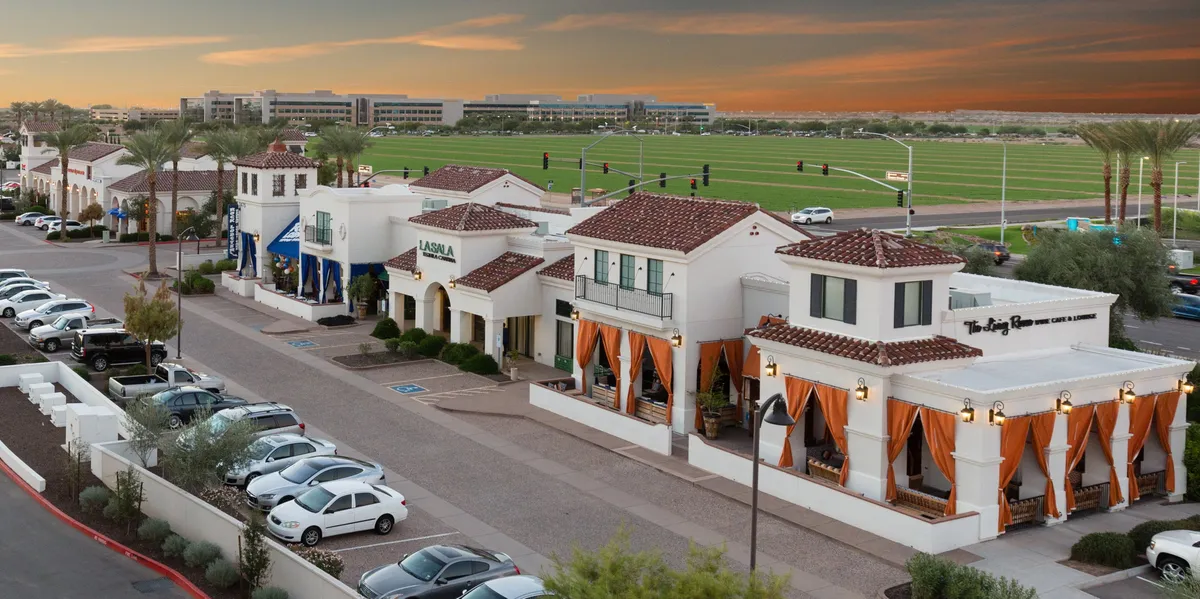 A lively exterior scene showcasing attractive restaurant facades with orange drapes and a green lawn area, inviting people for al fresco dining or evening strolls. The setting exudes a welcoming, walkable neighborhood atmosphere.