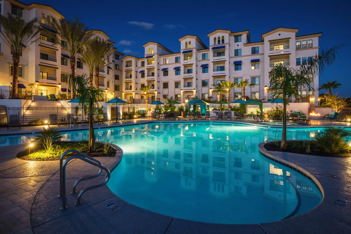 A tranquil outdoor swimming pool is surrounded by palm trees and comfortable seating areas. The pool area is illuminated with subtle lighting fixtures, and modern buildings form the backdrop, creating a resort-like ambiance.