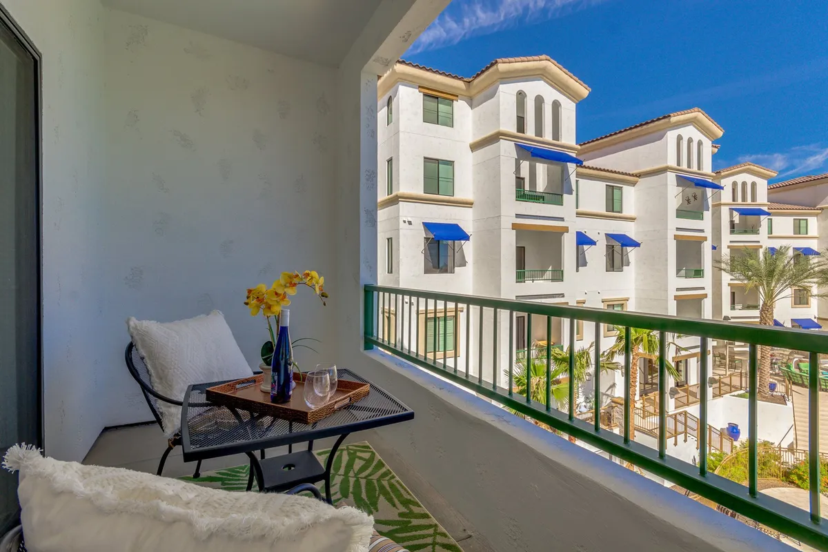 A serene balcony setup overlooks the sparkling pool area, featuring comfortable seating, a flower vase, and a panoramic view of the lush community grounds. The scene suggests relaxation and moments of leisure after a day on the golf course.